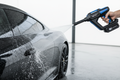 Portable cordless pressure washer, sleek black/blue design, spraying water on a car surface, clean white background and lifestyle setting, water droplets detailed, high-tech handheld washer, 8K marketing photo.